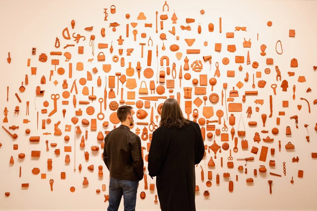 Two people view an intricate wall installation featuring a variety of red, ceramic-like objects in an art gallery.