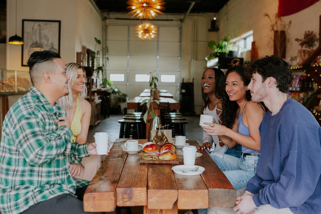A group of five friends enjoying coffee and pastries at a cozy café with a warm atmosphere and wooden furniture.