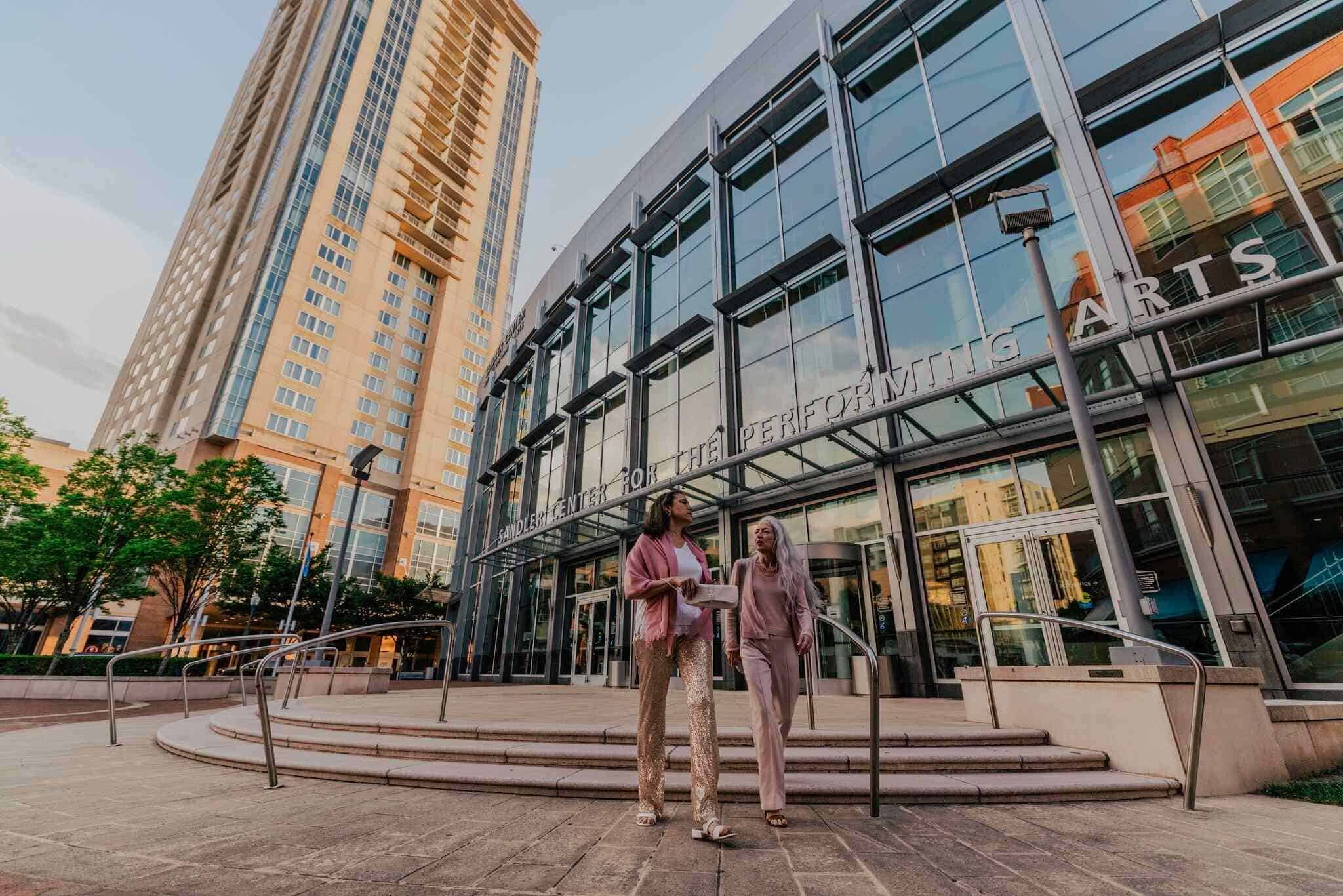 Two women, casually dressed, walk down the steps of the Sandler Center for the Performing Arts with a modern high-rise building in the background.