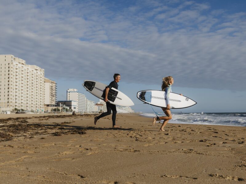 A man and a woman run along a sunny beach, each carrying a surfboard, with buildings and ocean waves in the background.