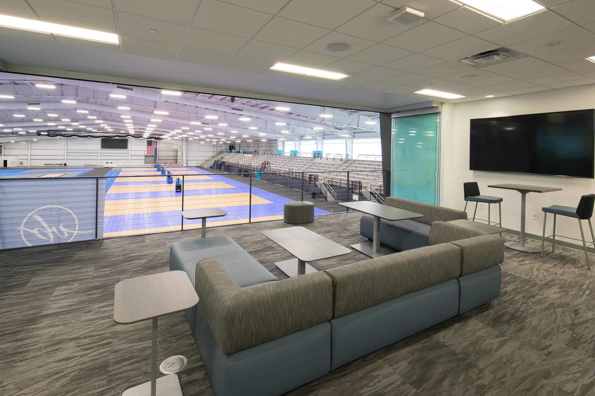 Modern lounge overlooking a sports arena with wooden floors, bleachers, and blue mats, featuring comfortable seating and tables.