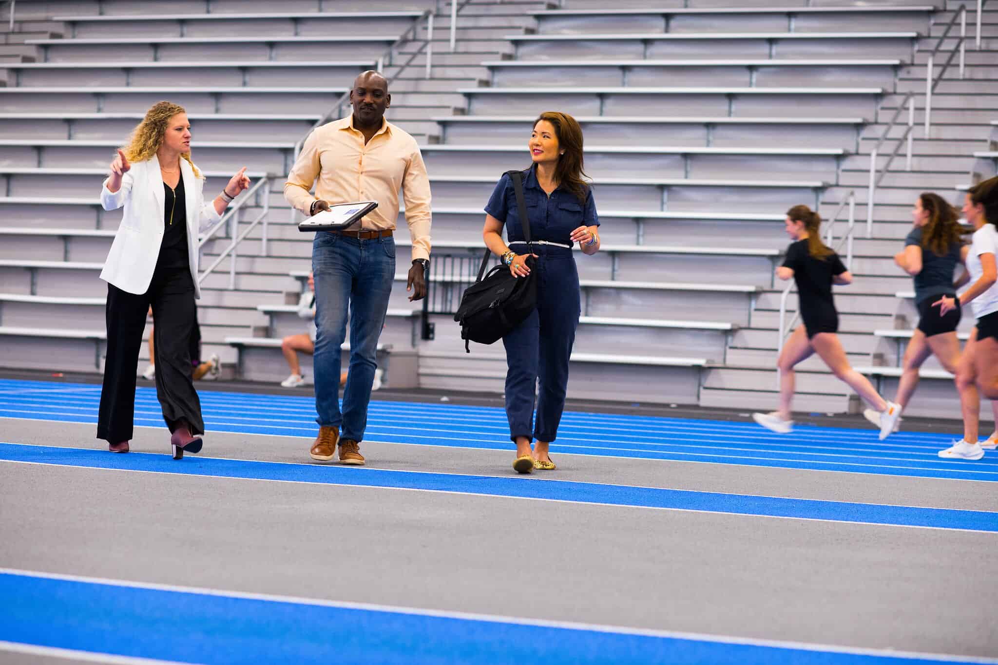 Three professionals walk on a blue track inside a sports facility, discussing plans as athletes practice in the background.