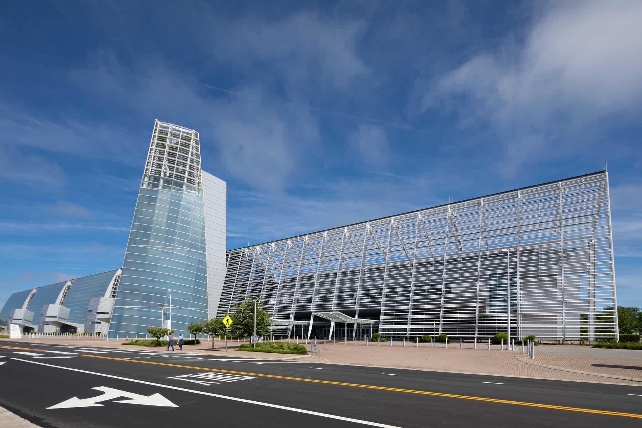 Modern architectural structure with glass and metal facade under a blue sky, featuring landscaped entrance and pedestrian area.