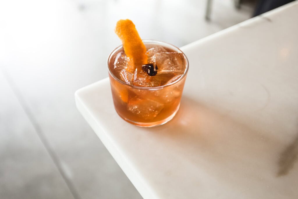A refreshing cocktail with ice, garnished with an orange peel, served in a clear glass on a marble countertop.