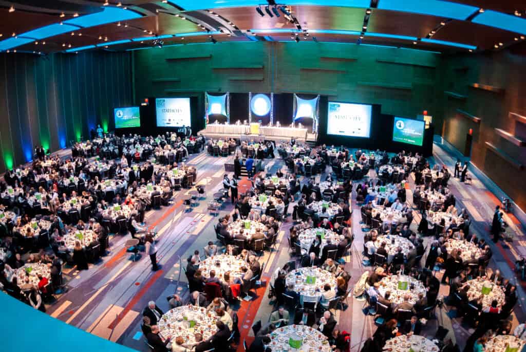 Aerial view of a large banquet hall filled with attendees seated around circular tables, with a stage and screens in the background.