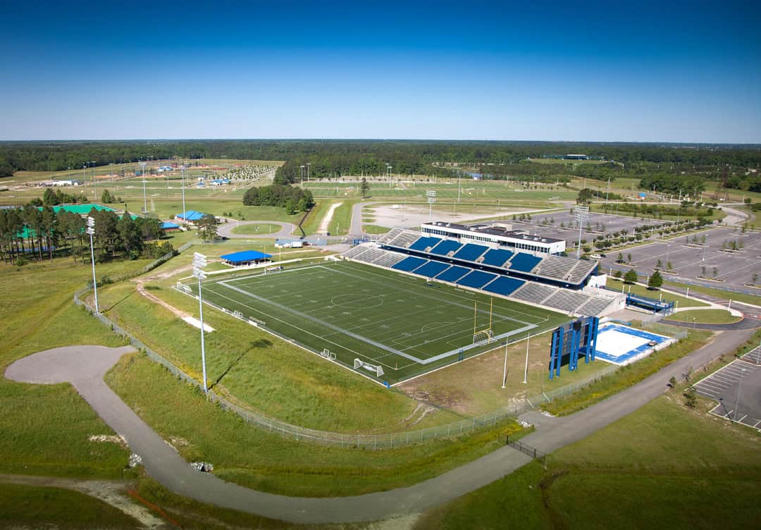 Aerial view of Princess Anne Sports Complex and Virginia Beach Sportsplex. A large sports field with seating and lighting, surrounded by green grass and a parking area in a recreational complex.