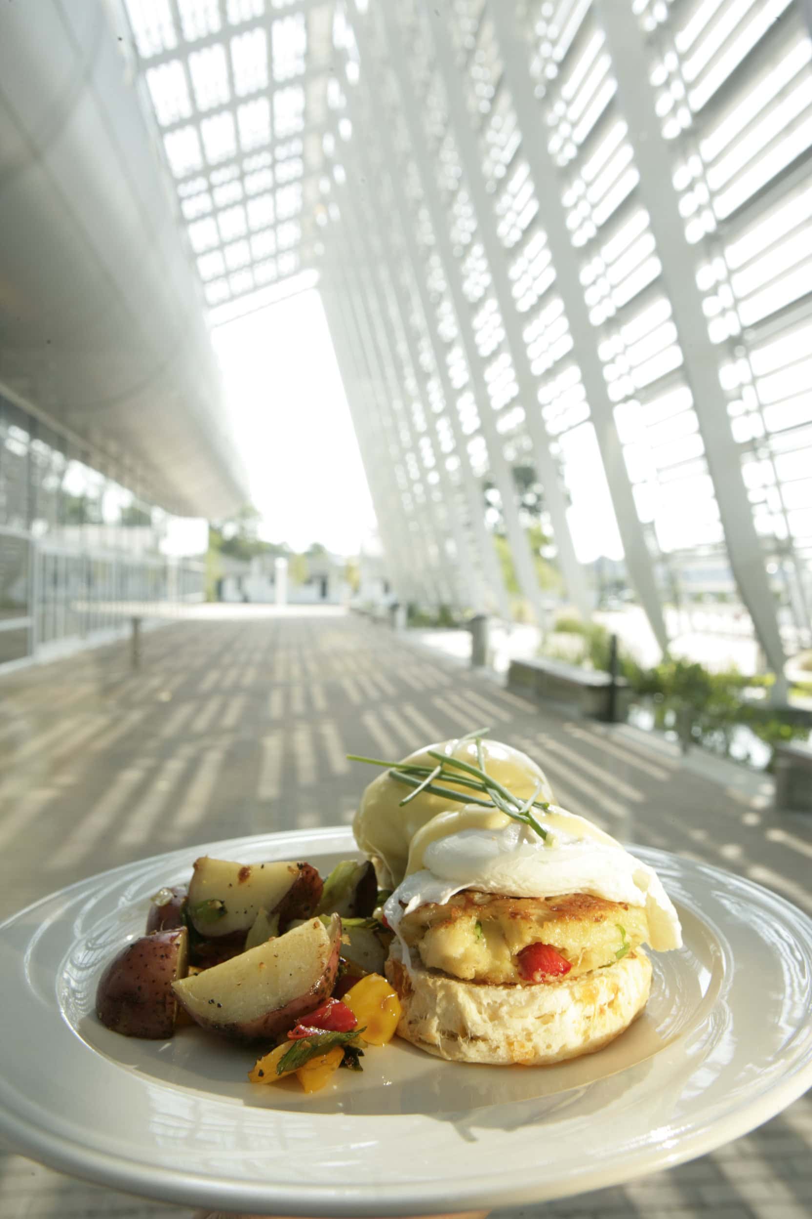 A plate of breakfast featuring a biscuit topped with poached eggs and hollandaise sauce, accompanied by seasoned roasted potatoes and colorful vegetables, set against a modern architectural backdrop with abundant natural light.