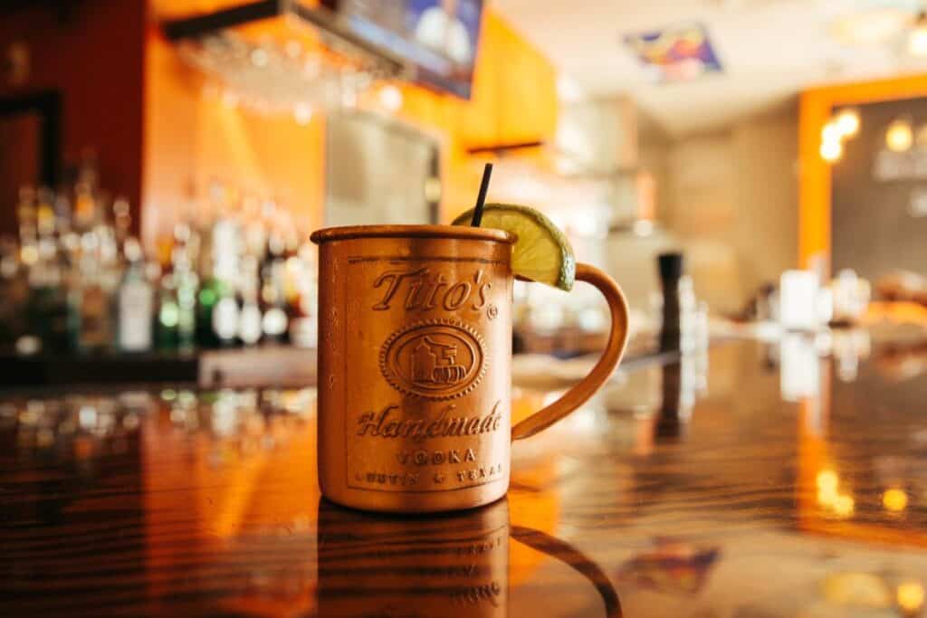 A close-up of a copper mug with Tito's Vodka branding and a lime wedge, set on a polished wooden bar with blurred bottles in the background.