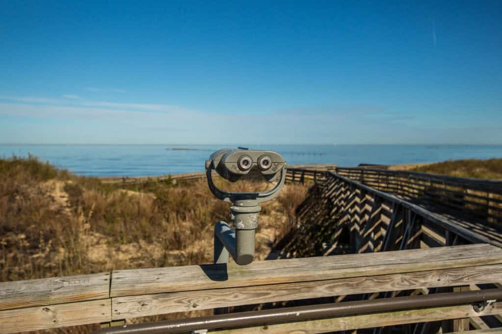 A binocular viewer on a wooden deck overlooking a calm ocean and a clear blue sky.