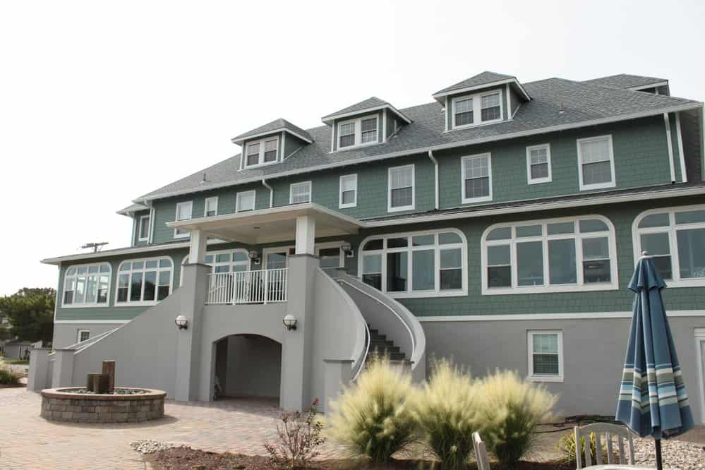 Historic green beachfront hotel with expansive windows, circular staircase, and landscaped patio featuring seating and plants.