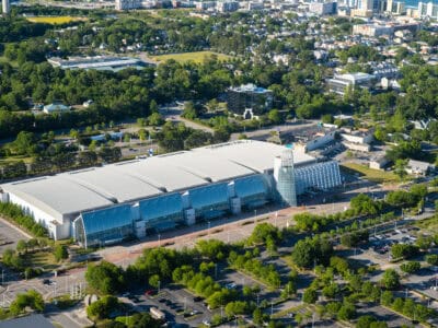 Aerial view of a modern convention center surrounded by green trees and parking lots, showcasing its glass facade and architecture.