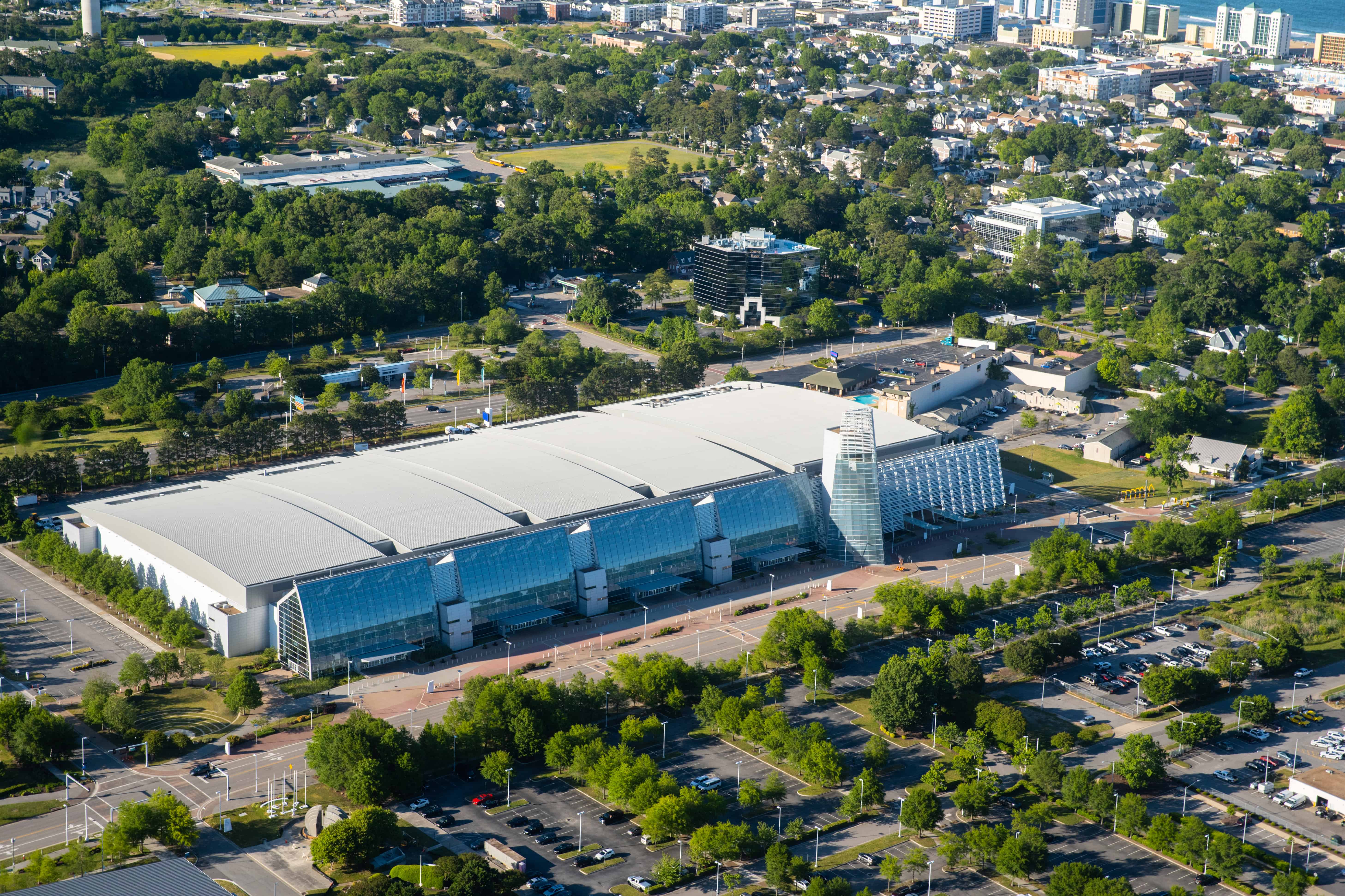 Aerial view of the modern Virginia Beach Convention Center surrounded by green trees and parking lots, showcasing its glass facade and architecture.