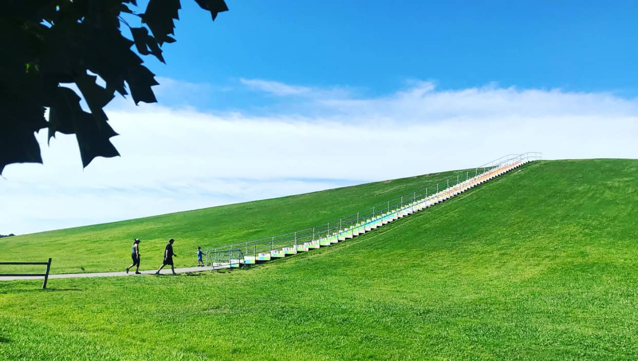 A scenic view of a colorful staircase leading up a grassy hill, with people walking along a pathway under a blue sky.