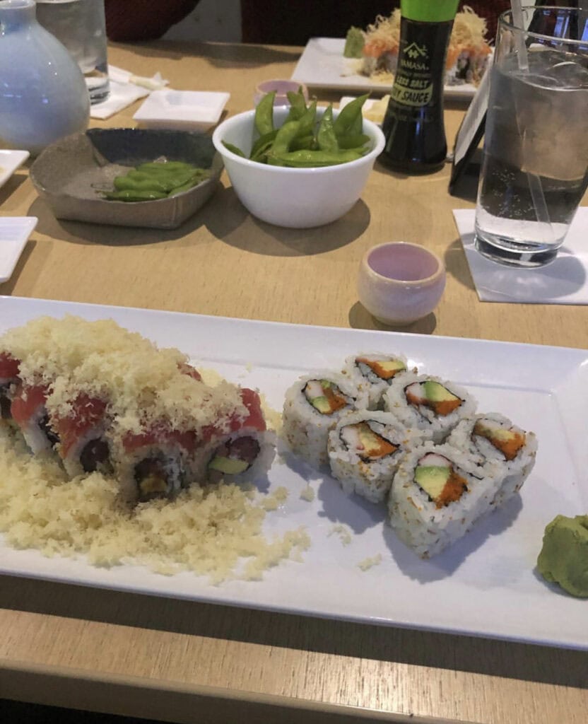 A sushi platter featuring a colorful roll topped with crispy flakes, alongside California rolls, with edamame and soy sauce in the background.