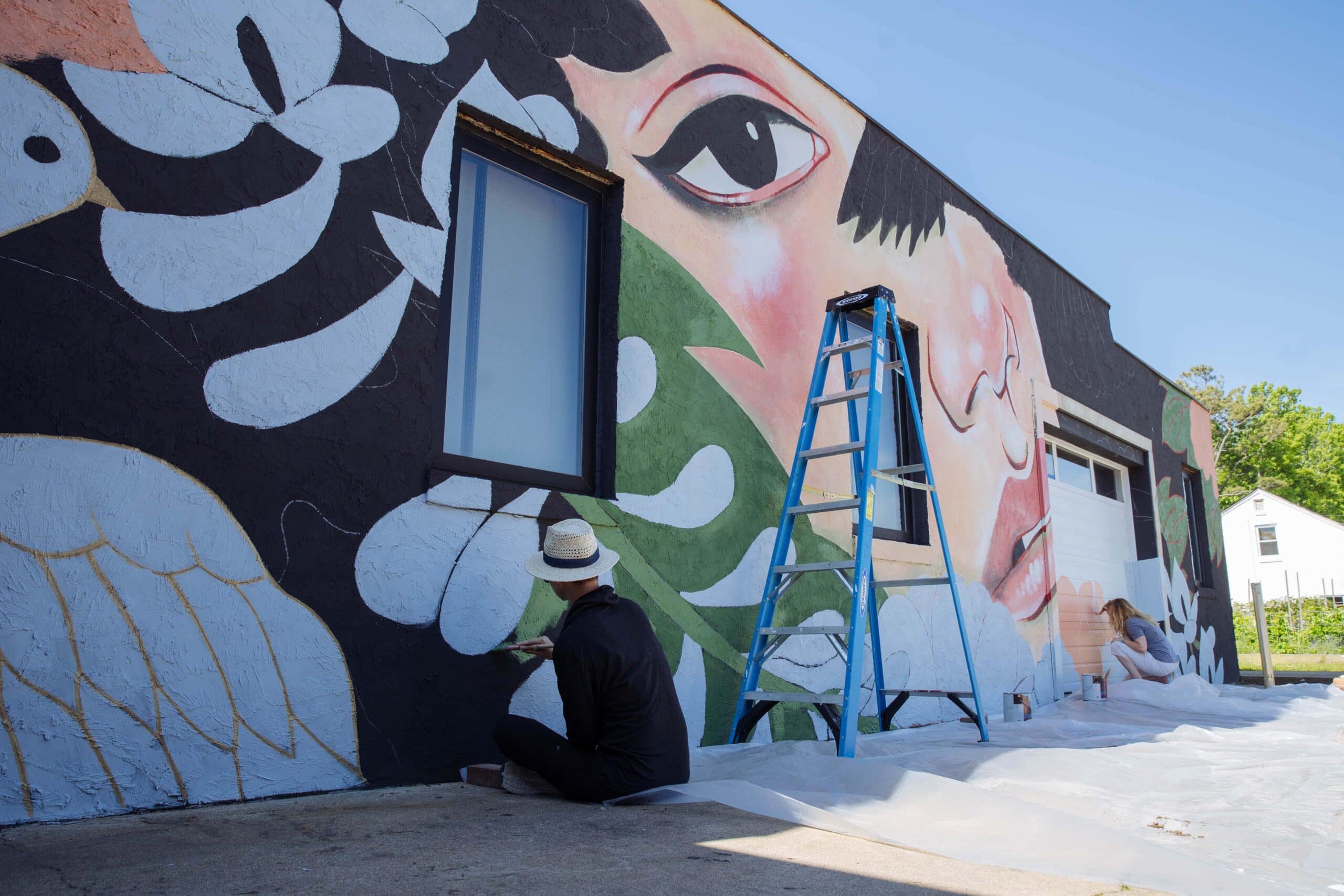 A mural artist works on a colorful wall mural at Esoteric featuring a face and leaves, with a ladder and paint supplies nearby.