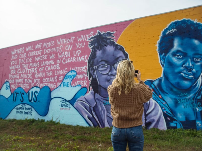 A person with long blonde hair takes a photo in front of a colorful mural in the ViBe Creative District featuring two portraits and poetic text about water and opportunity.