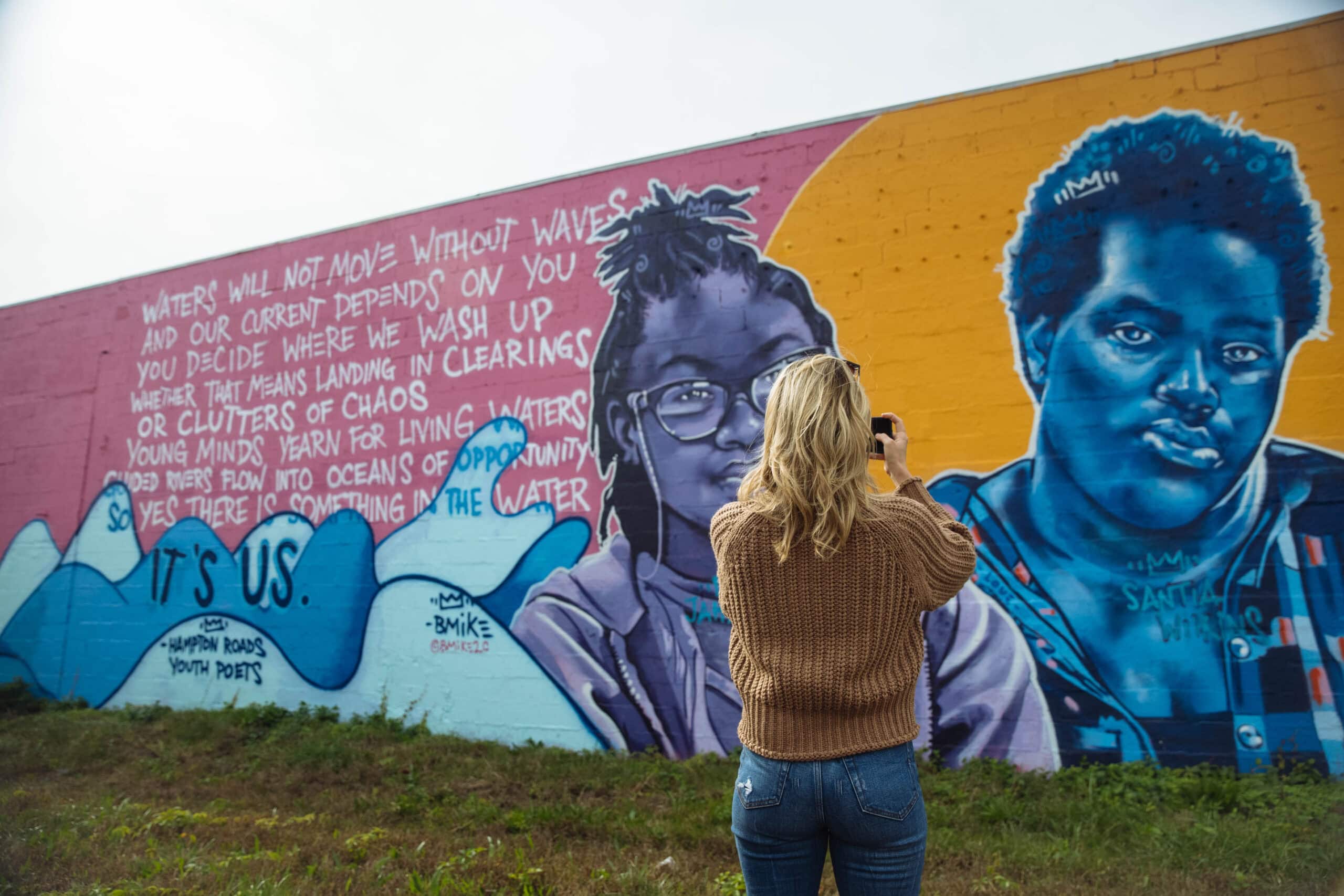 A person with long blonde hair takes a photo in front of a colorful mural in the ViBe Creative District featuring two portraits and poetic text about water and opportunity.
