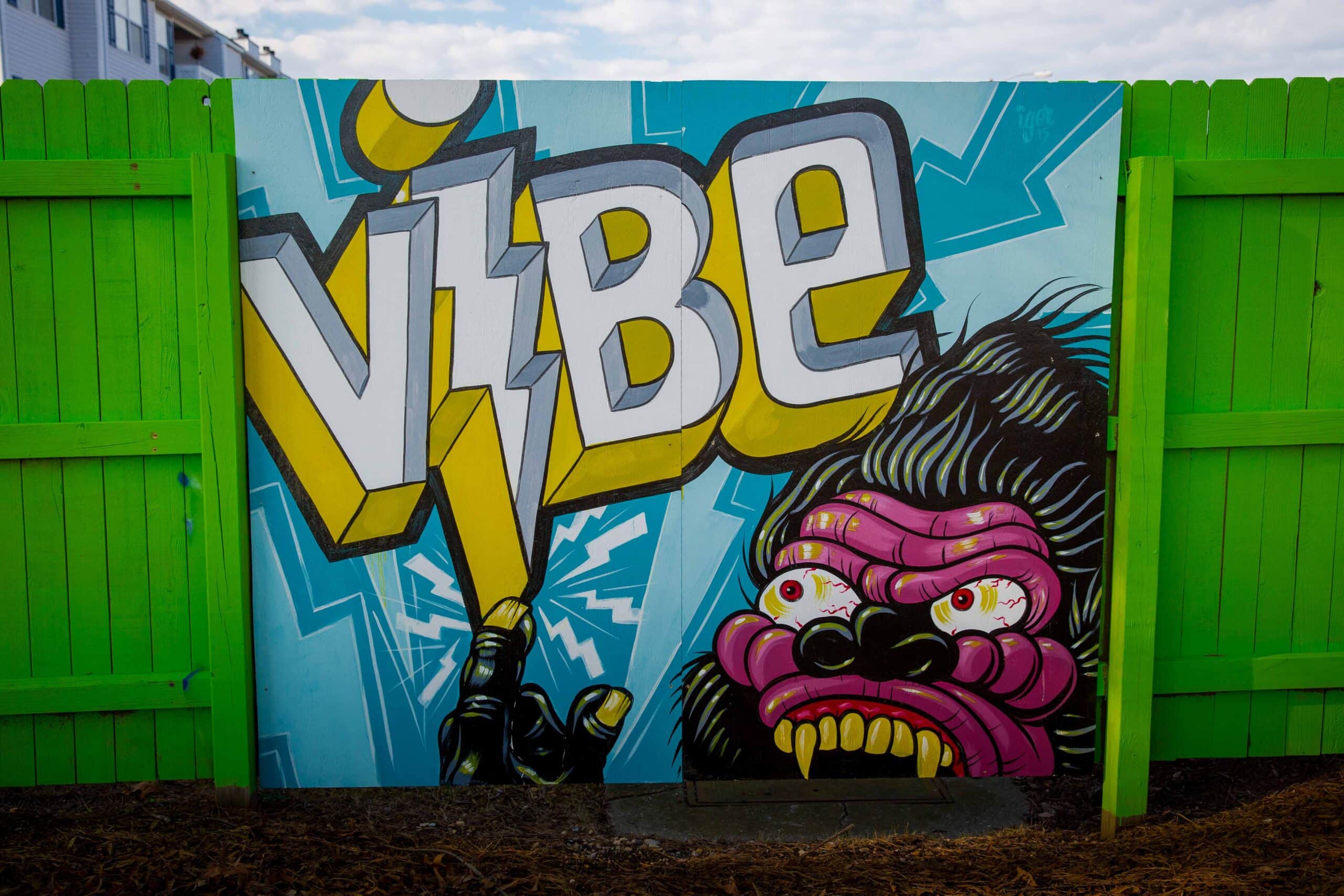 A vibrant mural featuring the word "VIBE" in bold yellow, alongside a colorful, fierce gorilla face with exaggerated features, set against a blue background, all framed by a bright green fence in the ViBe Creative District.