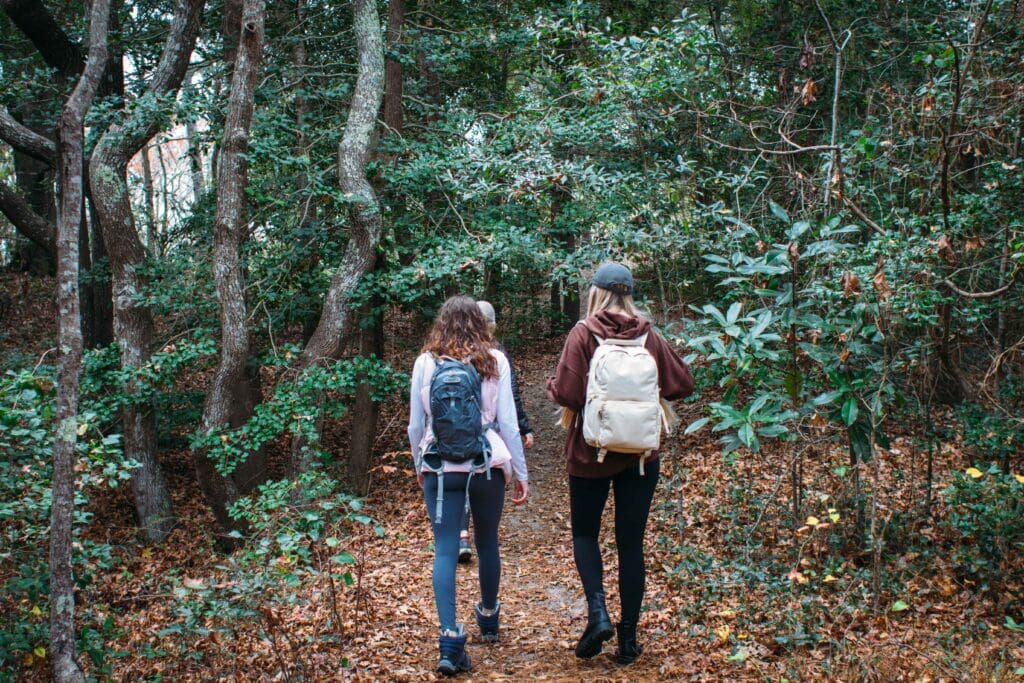 Three women walking along a wooded trail with backpacks, surrounded by a forest setting at First Landing State Park.