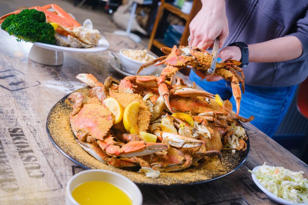 A close-up of a person preparing a platter of seasoned crabs garnished with lemon, alongside sides of butter and coleslaw at Margie & Ray's Crabhouse and Restaurant.