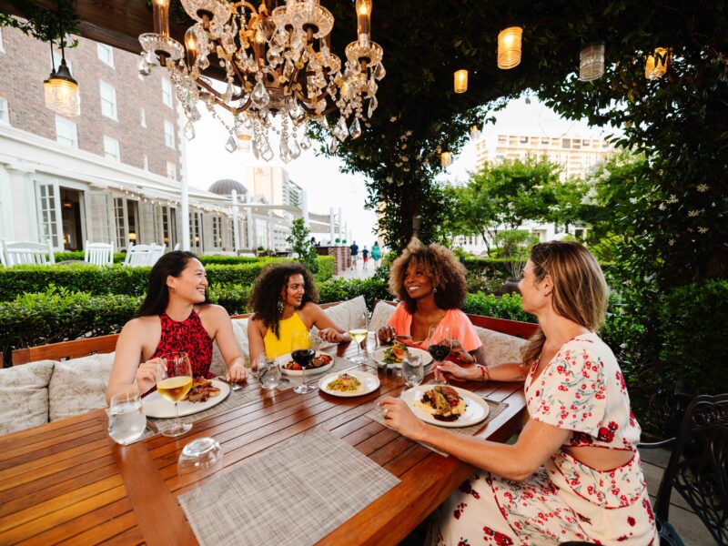 A group of four women enjoying a meal outdoors under a chandelier, with plates of food and drinks on a wooden table amid greenery during Summer.