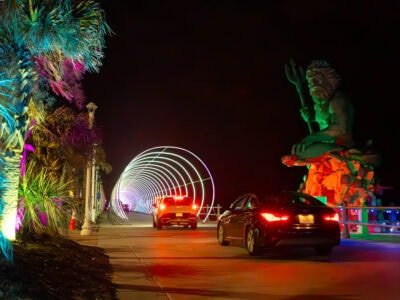 A vibrant nighttime scene with colorful lights illuminating palm trees, cars driving through a glowing arch, and a large statue of King Neptune by the Virginia Beach Boardwalk.