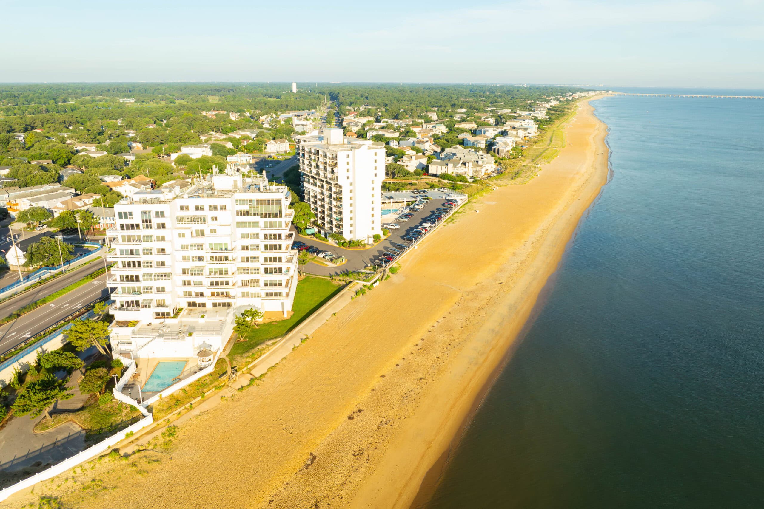 Aerial view of a sandy beach lined with coastal buildings, showcasing lush greenery and calm waters in the distance at Chic's Beach in Virginia Beach.