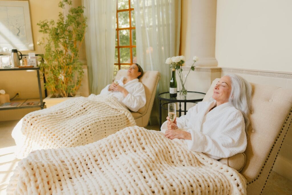 Two women relax in plush chairs at spa, wrapped in chunky knit blankets, each holding a glass of sparkling drink at Flowering Almond Spa in The Founders Inn.