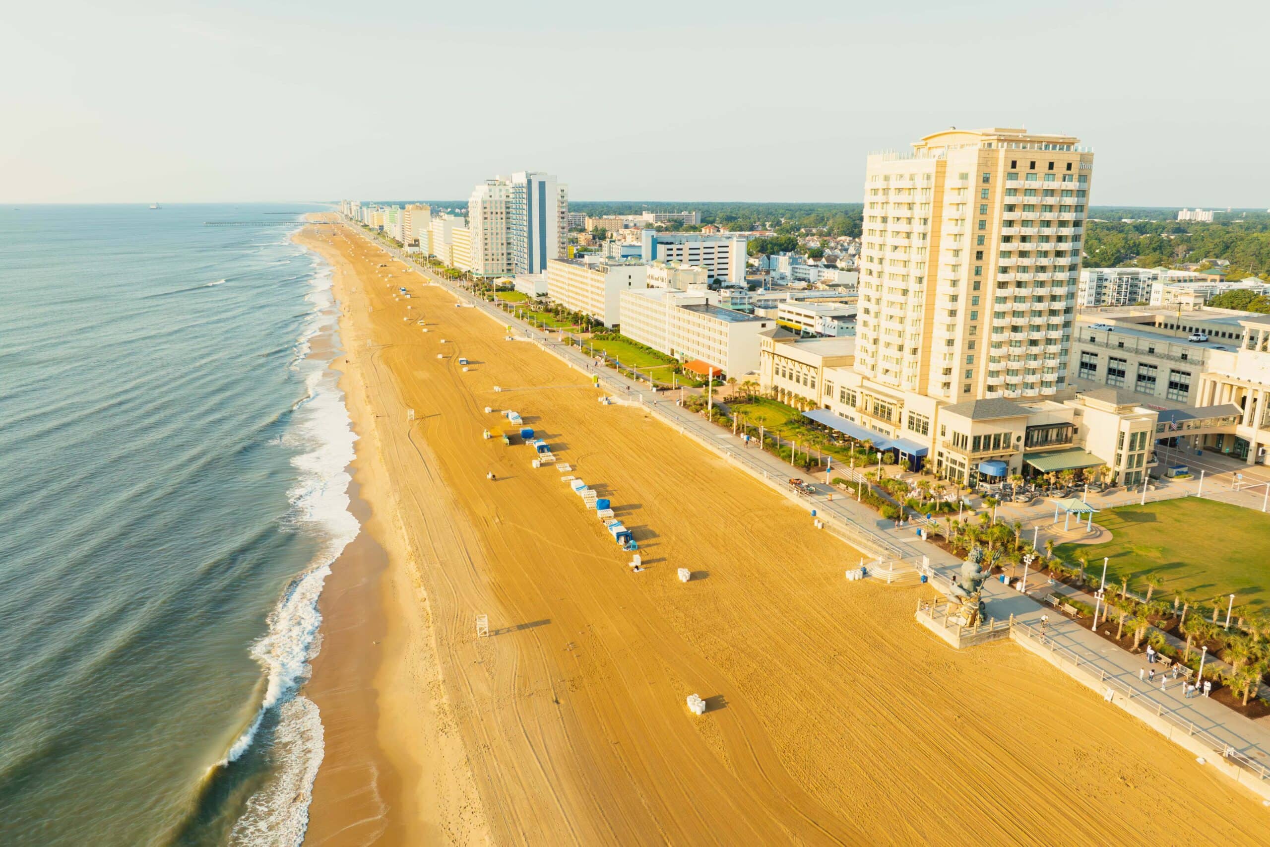 Aerial view of a sandy beach with ocean waves, lined by hotels and a boardwalk in Virginia Beach.