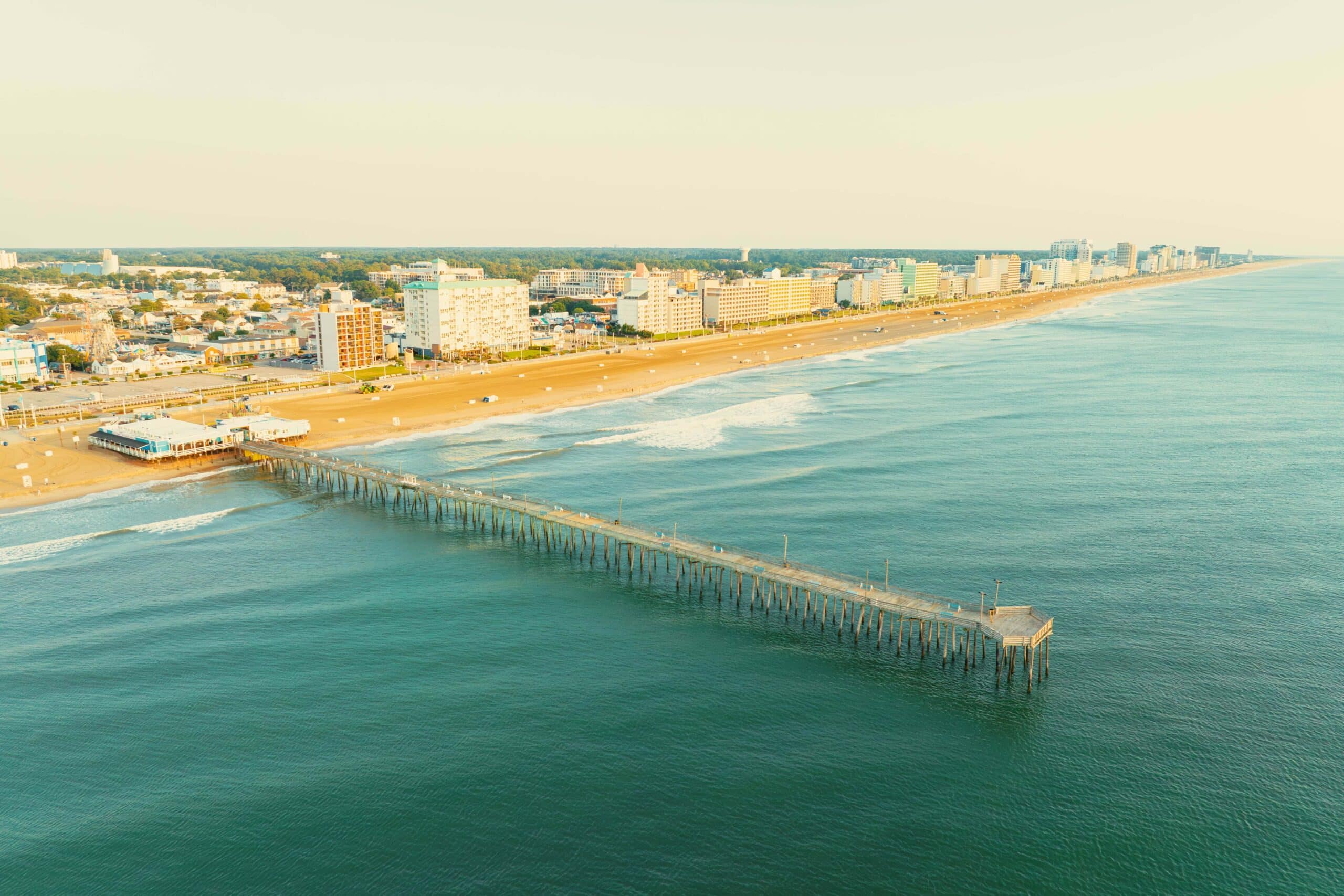 Aerial view of Virginia Beach Fishing Pier extending into calm waters, with sandy beach and beachfront hotels in the background at the Oceanfront.