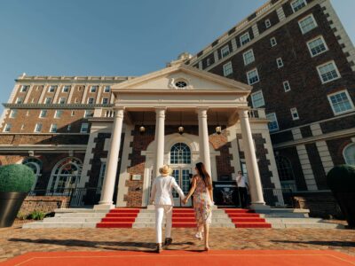 A couple dressed elegantly is walking hand in hand on a red carpet towards The Historic Cavalier with columns and a manicured landscape.