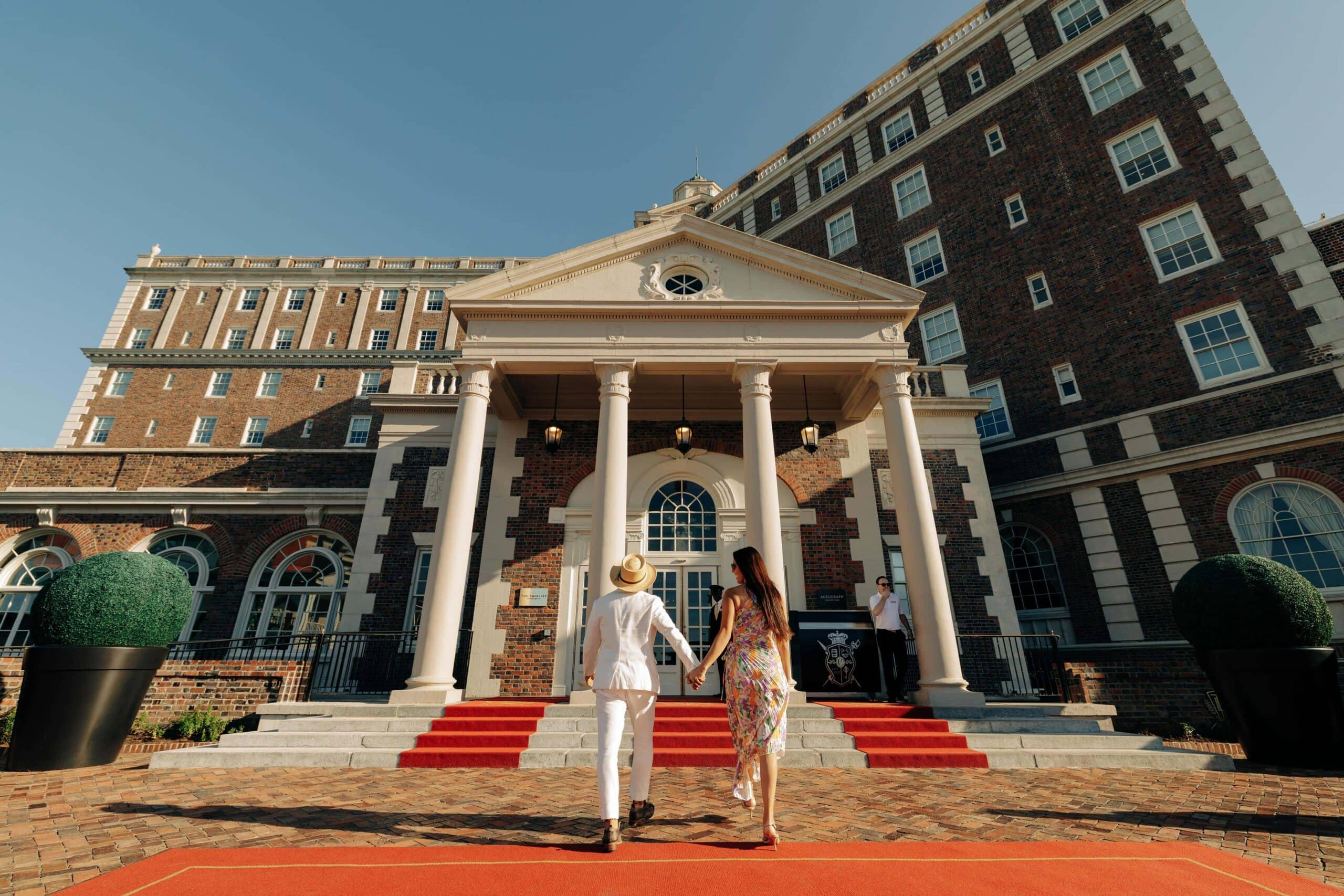 A couple dressed elegantly is walking hand in hand on a red carpet towards The Historic Cavalier with columns and a manicured landscape.