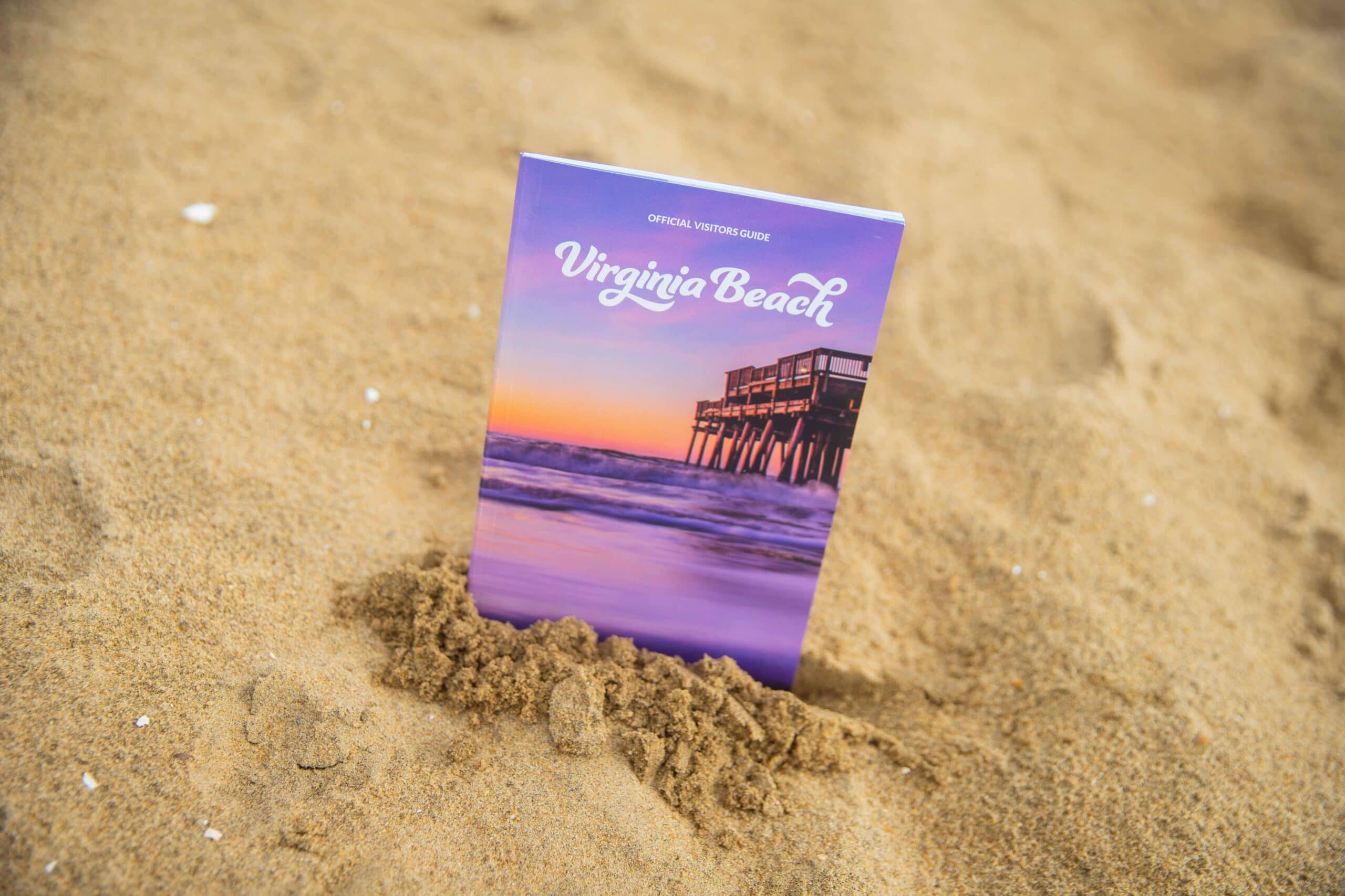 A Virginia Beach official visitors guide partially buried in sand, featuring a colorful cover with a pier at sunset in.