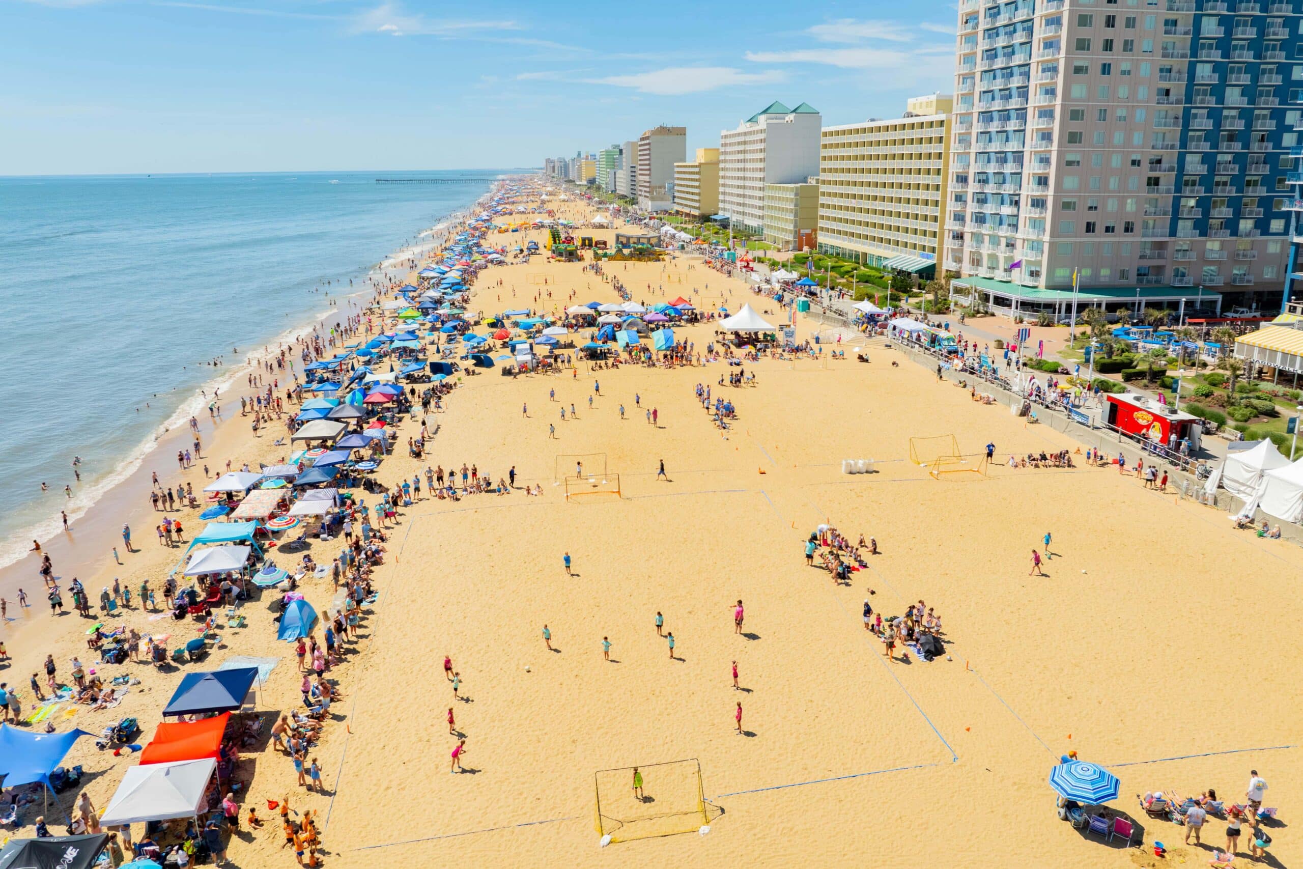 Aerial view of a busy beach with colorful umbrellas, people enjoying sand activities, and oceanfront hotels along the coastline.