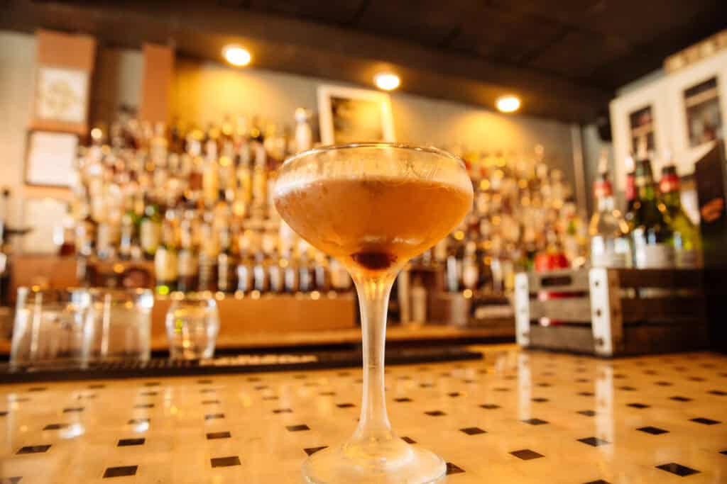 A close-up of a cocktail in a glass with a cherry, set against a blurred bar filled with bottles.