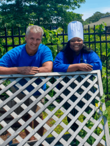 Two individuals, a man in a blue shirt and a woman in a blue chef jacket and white chef hat, smile as they pose together behind a white lattice fence in a green garden setting.