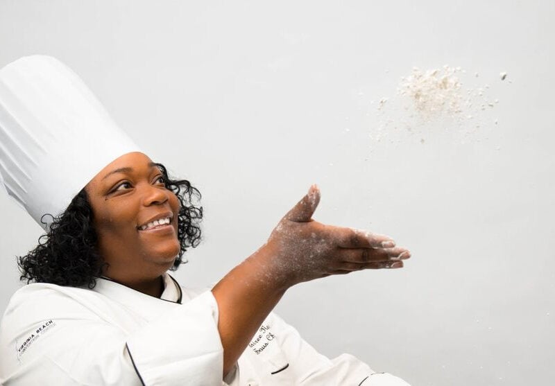 A smiling female chef in a white uniform and tall hat playfully tosses flour into the air against a light background.