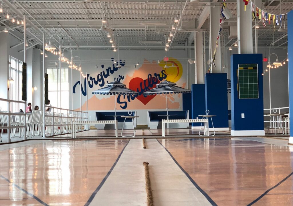 Beachside Social is pictured with a gymnasium type space with striped blue and white umbrellas and overhead lighting. It is located by the Virginia Beach Oceanfront.