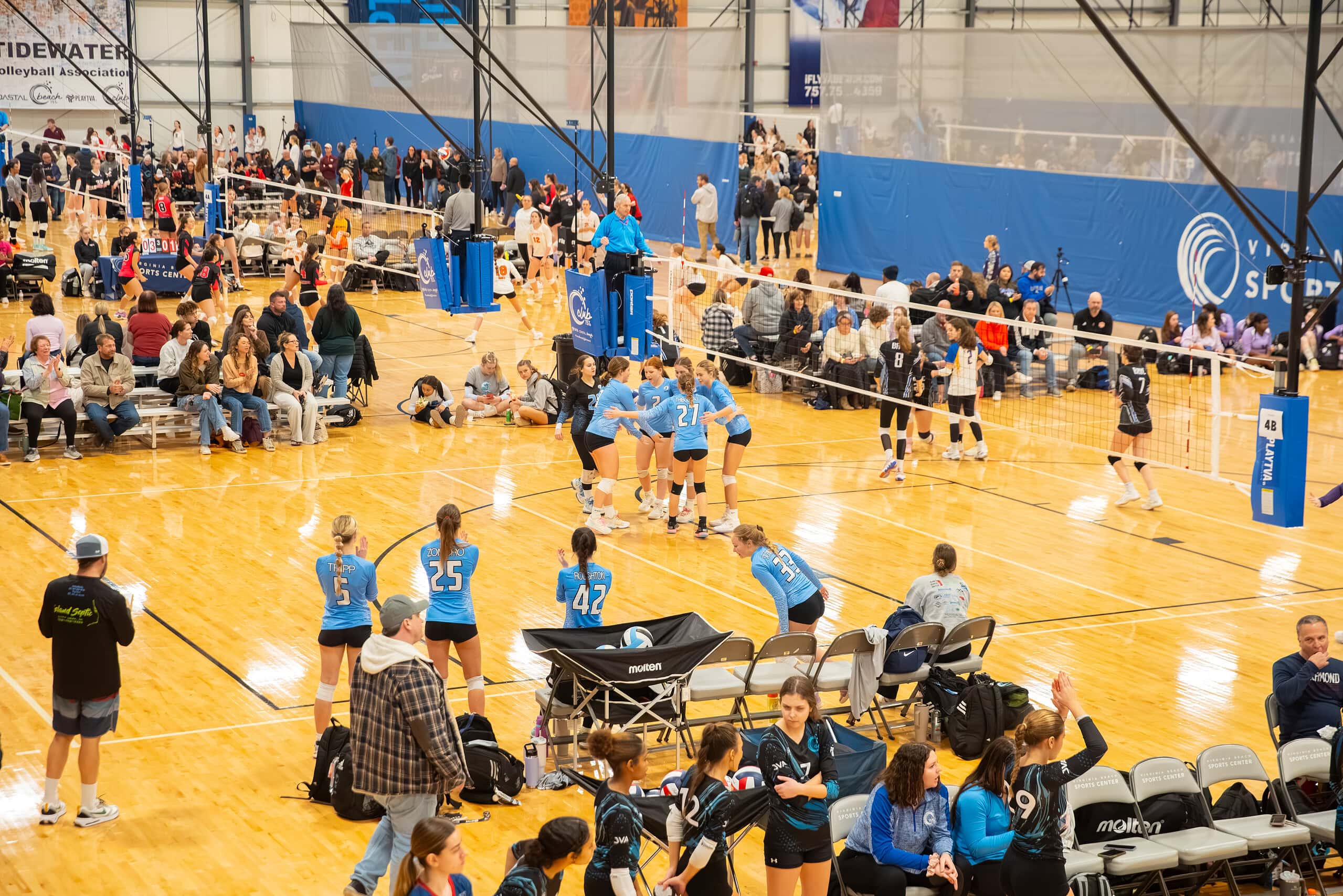 A female volleyball team huddling during a match surrounded by an excited crowd with other volleyball matches taking place in the background at the Virginia Beach Sports Center.