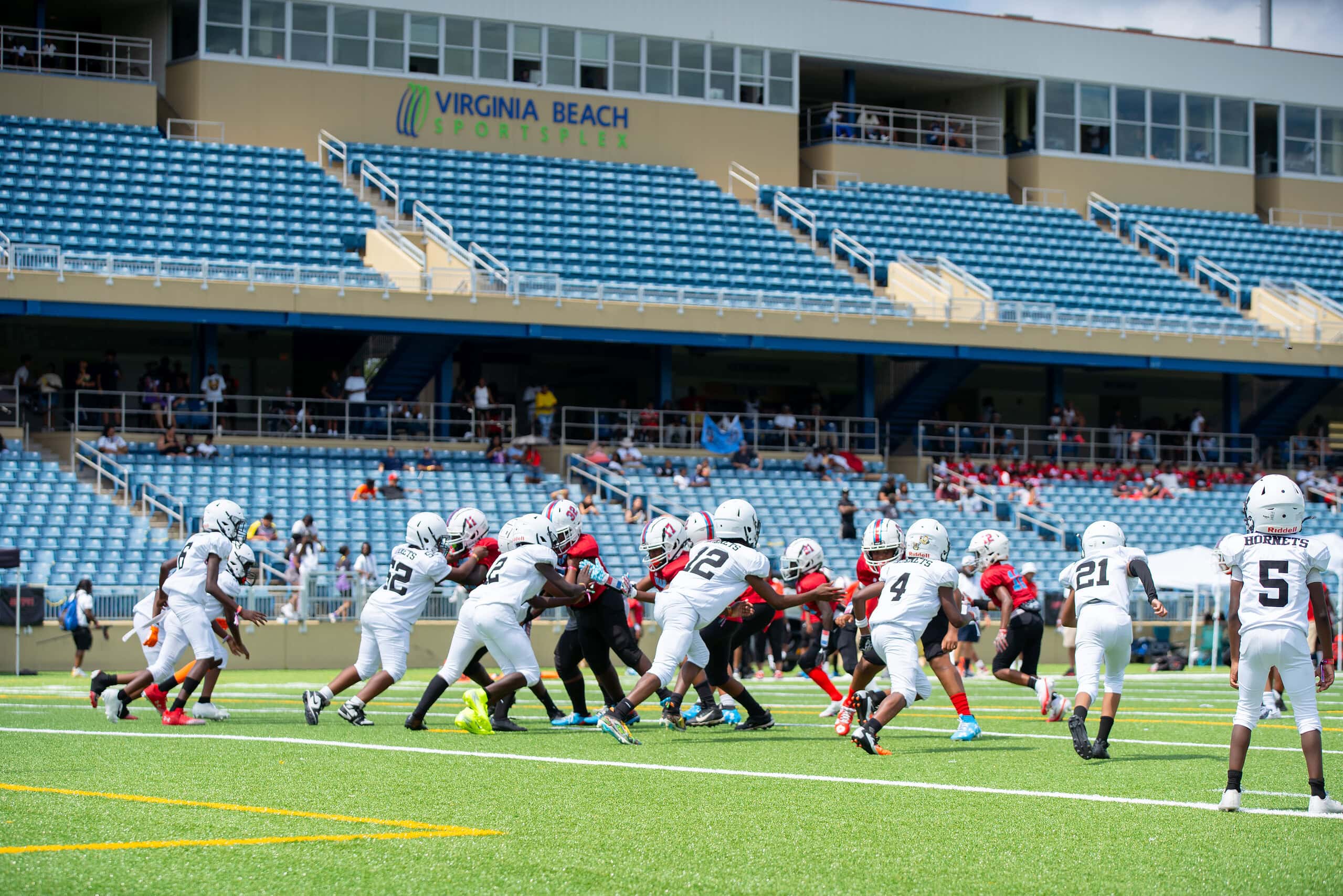 Energetic young football players competing on a turf field with a crowd on-looking in the background in stadium seating at Virginia Beach Sportsplex.