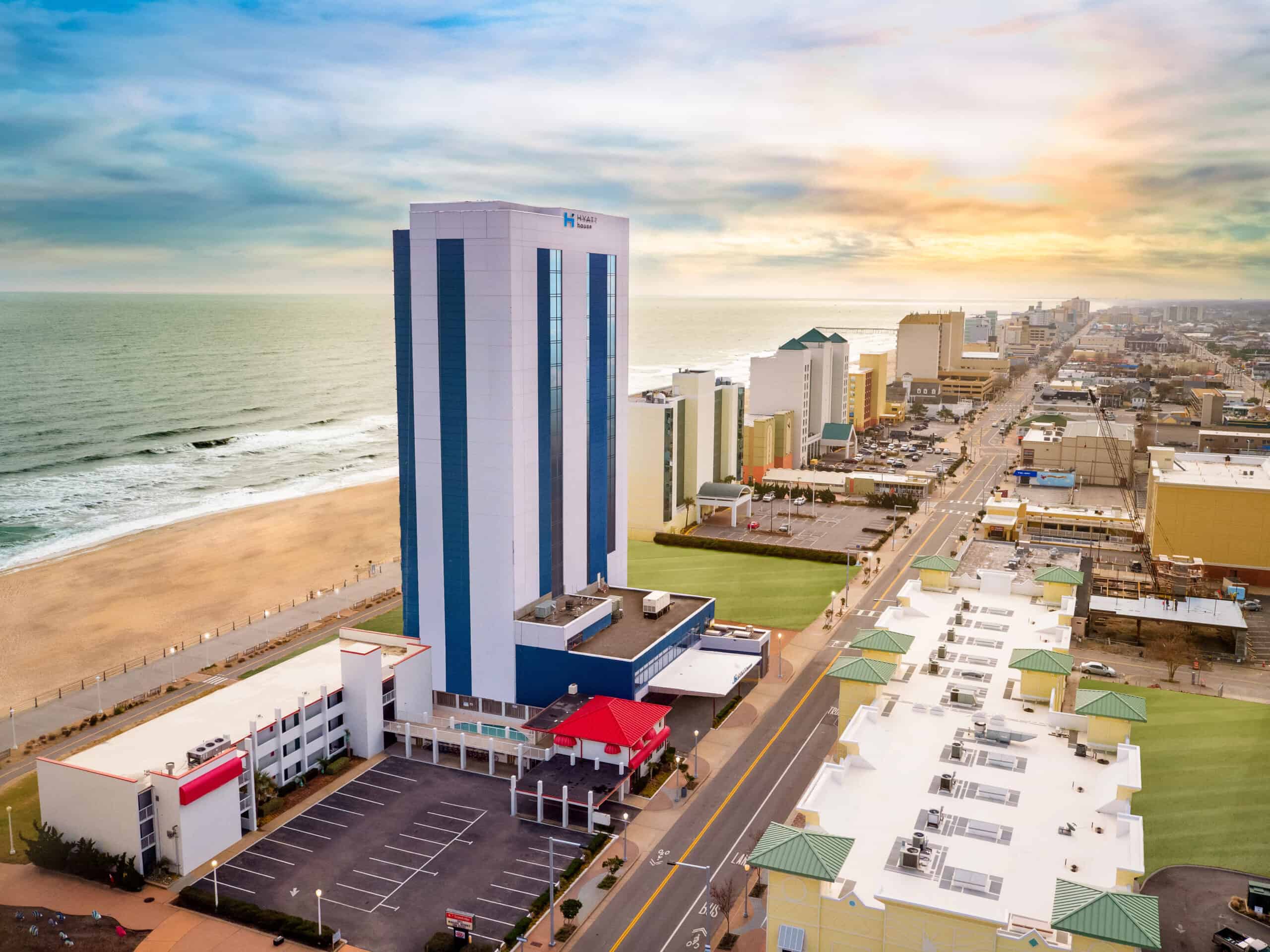 Aerial view of a beachside hotel and coastline, featuring ocean waves, sandy beach, and a bustling boardwalk area.