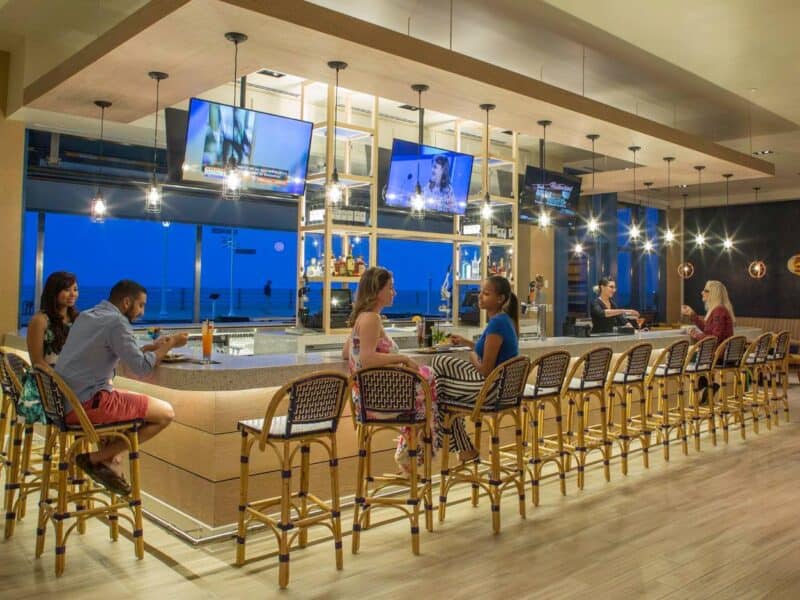 A modern bar scene featuring patrons enjoying drinks and food, with a view of the ocean at twilight and multiple TVs displaying content.