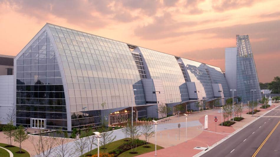 Modern glass building with an angular design, surrounded by landscaped grounds and a road, under a colorful sky at dusk.
