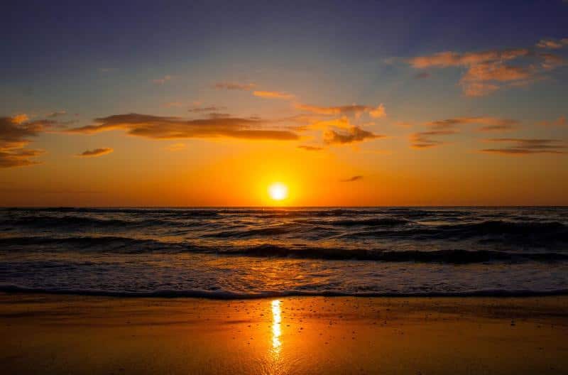Sunset over the ocean with an orange sky, scattered clouds, and waves gently lapping at the shore.