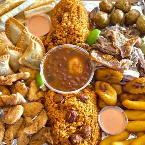 A platter with rice, beans, fried plantains, fried chicken, empanadas, pork, lime wedges, and sauce cups.