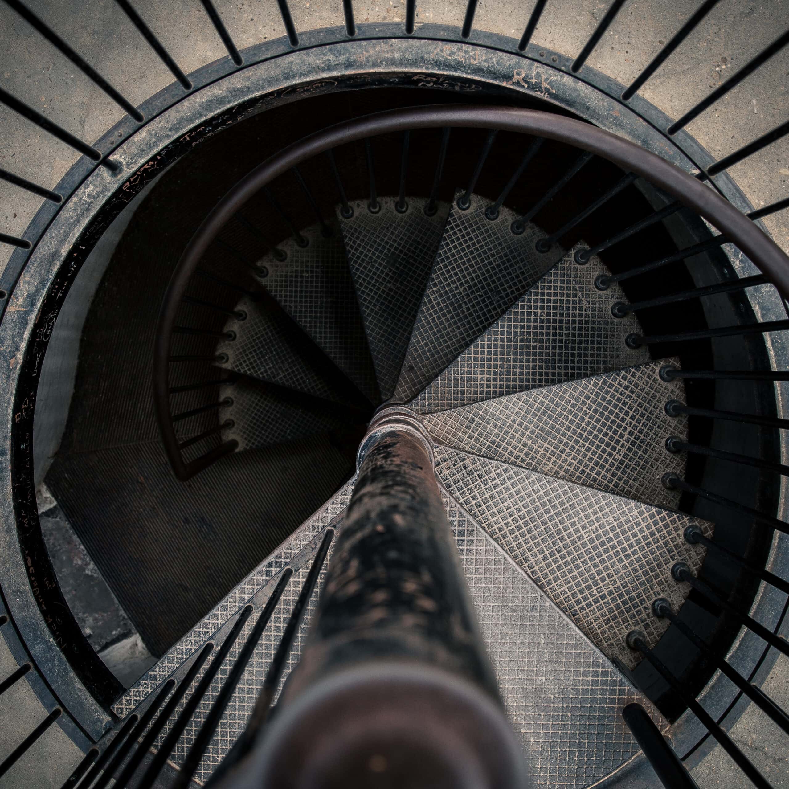 Spiral staircase with metal railing and textured steps, viewed from above, creating a circular pattern descending into a dimly lit area.