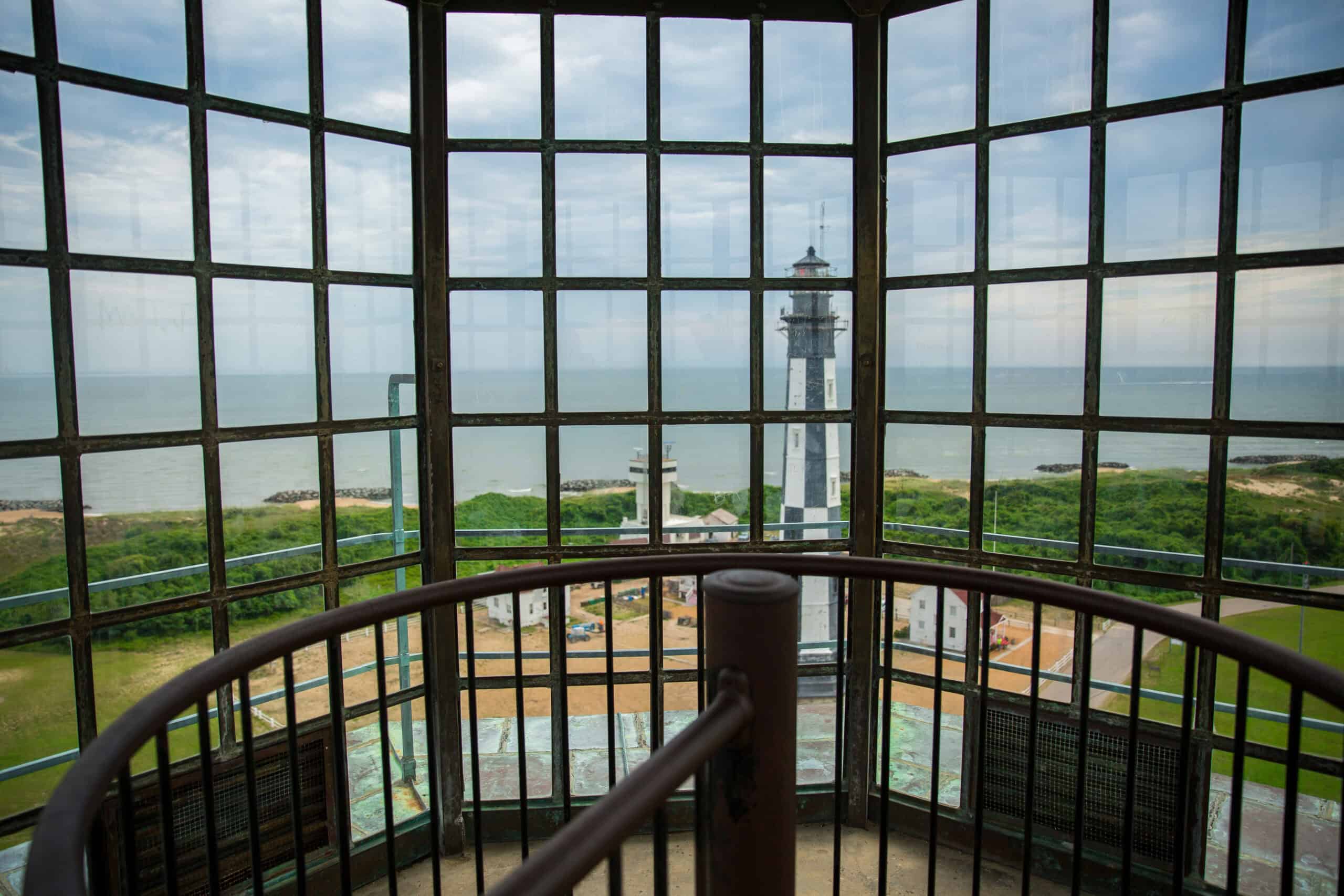 View from inside a lighthouse with large windows, overlooking a coastal landscape with another lighthouse, greenery, and the ocean under a cloudy sky.
