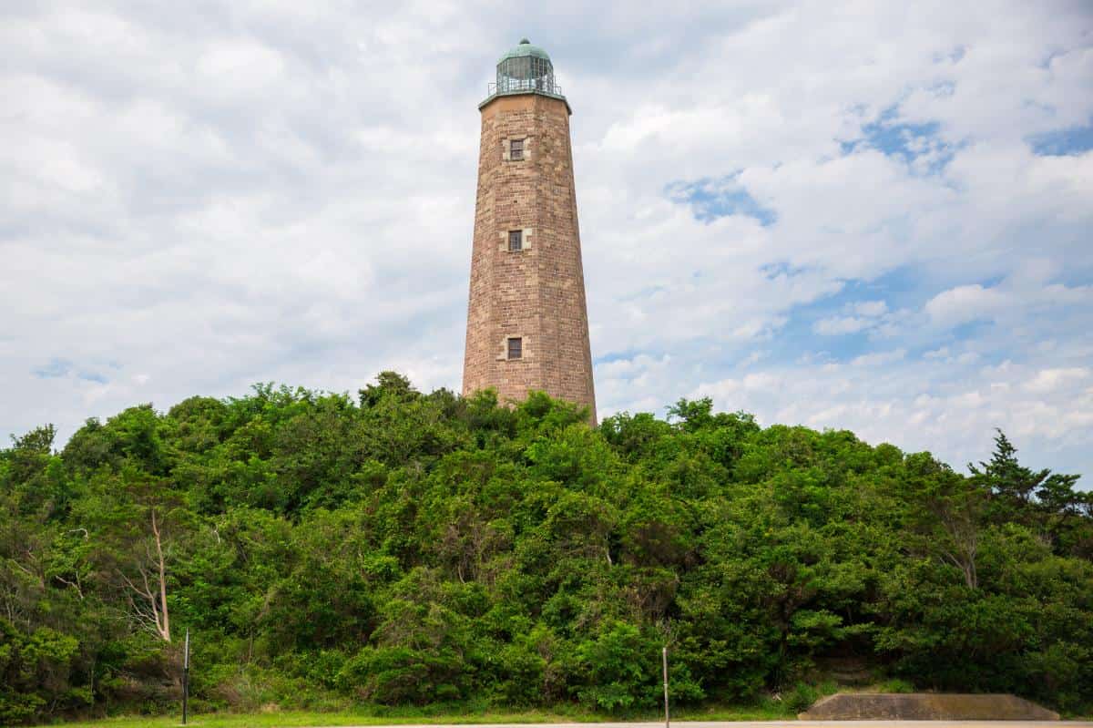 The old Cape Henry Lighthouse is a tall, brown stone structure which stands prominently atop a lush, green hill with a partly cloudy sky in the background.