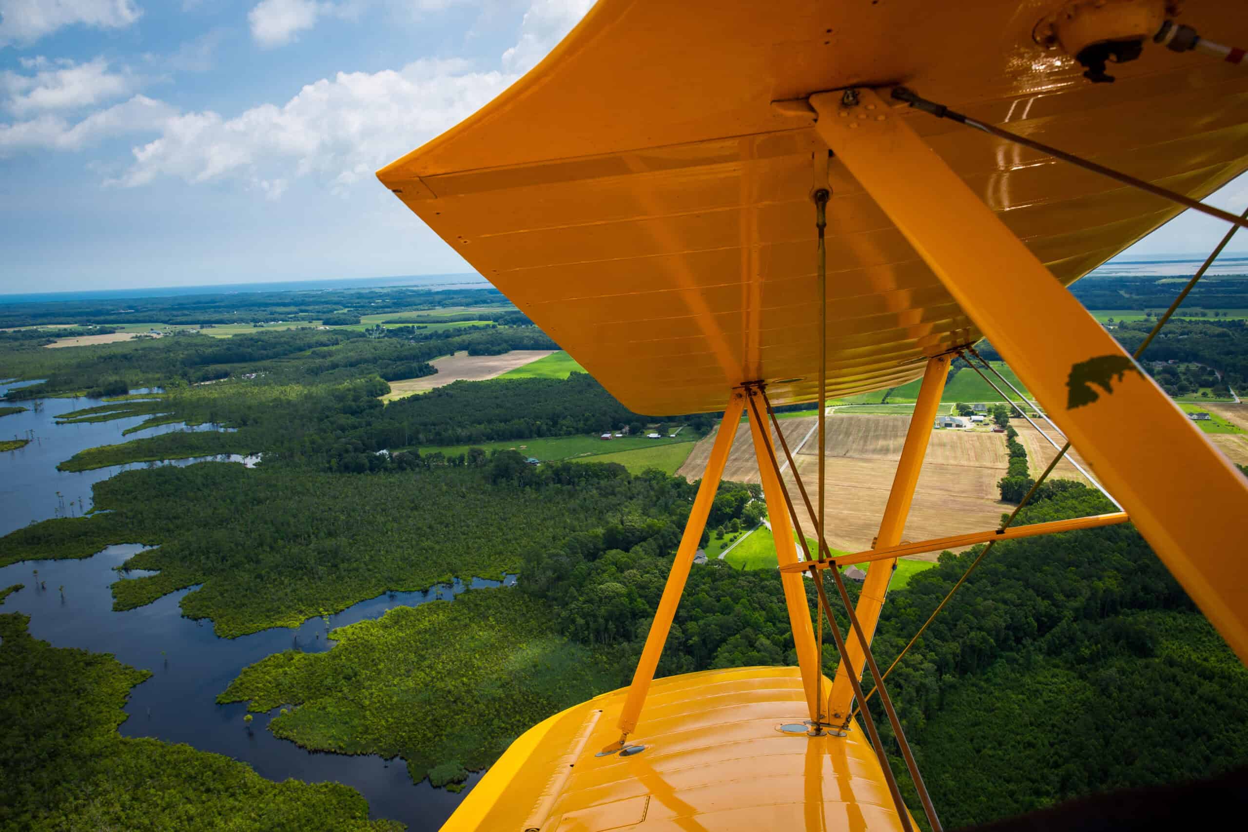 View from a yellow biplane flying over a lush, green landscape with rivers, fields, and forests under a partly cloudy sky.