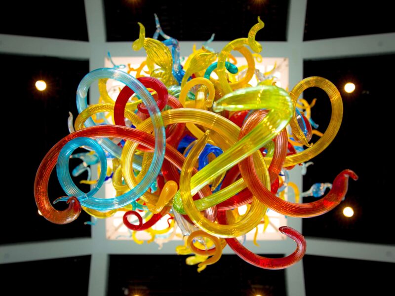 Colorful glass sculpture with intertwined red, yellow, and blue curves, viewed from below, set against a dark ceiling with lights.